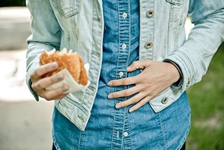 Frau in blauem Jeans Hemd und Jeans Jacke hält einen Burger in der Hand und hält sich den Bauch mit der anderen Frau in blauem Jeans Hemd und Jeans Jacke hält einen Burger in der Hand und hält sich den Bauch mit der anderen