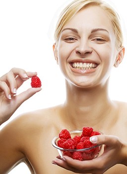 Junge blonde Frau lacht und hält eine Glasschüssel mit Himbeeren in der Hand und eine einzelne Frucht in der anderen