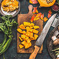 Holzbrett mit geschnittenem orangen Kürbis und Messer darauf umgeben von Lebensmittel