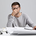 Ein Mann im grauen Pullover sitzt am Schreibtisch und reibt seine Augen unter der Brille