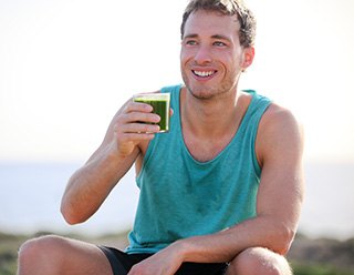 Mann in türkisfarbenem Tanktop sitzt in der Natur und hat ein Glas mit grünem Saft in der Hand