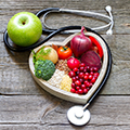 Obst und Gemüse in einer herzförmigen Schale, daneben liegen ein Apfel und ein Stethoskop  