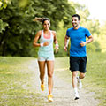 Ein Mann und eine Frau joggen auf einem Feldweg, im Hintergrund ist ein Wald zu sehen