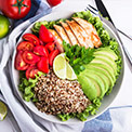 Weiße Schüssel mit Avocado, Hühnerfleisch, Quinoa, Tomaten und Salat auf einem weiß-blauen Geschirrtuch